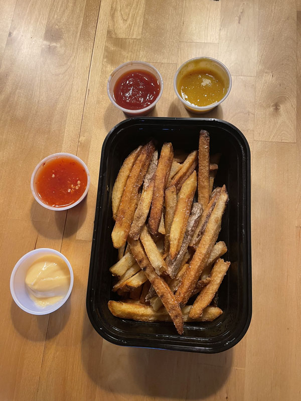 A plastic container filled with french fries. Surrounding the container of fries are four different plastic cups containing dipping sauces: Ketchup, Mayonnaise, Mustard, and an unknown spicy sauce. 