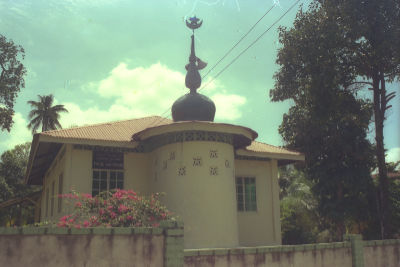 A photo of Masjid Wak Sumang showing its view from Punggol Road. The mosque has a hip roof and a cylindrical minaret extension topped with the star and crescent symbol of Islam. A low wall in front of the mosque separates it from the road.