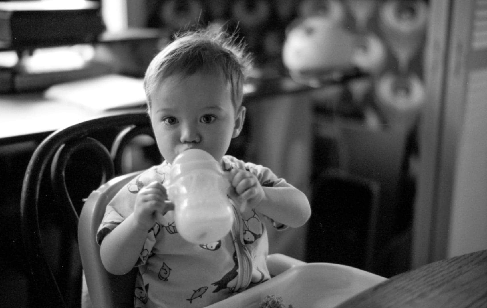 A baby drinking milk from a sippy cup at a dining room table