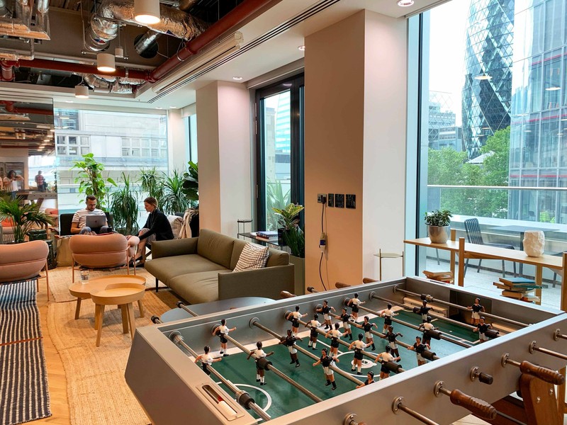 Employees work in a break room with a foosball table, comfy couches, and large windows looking out on a business district