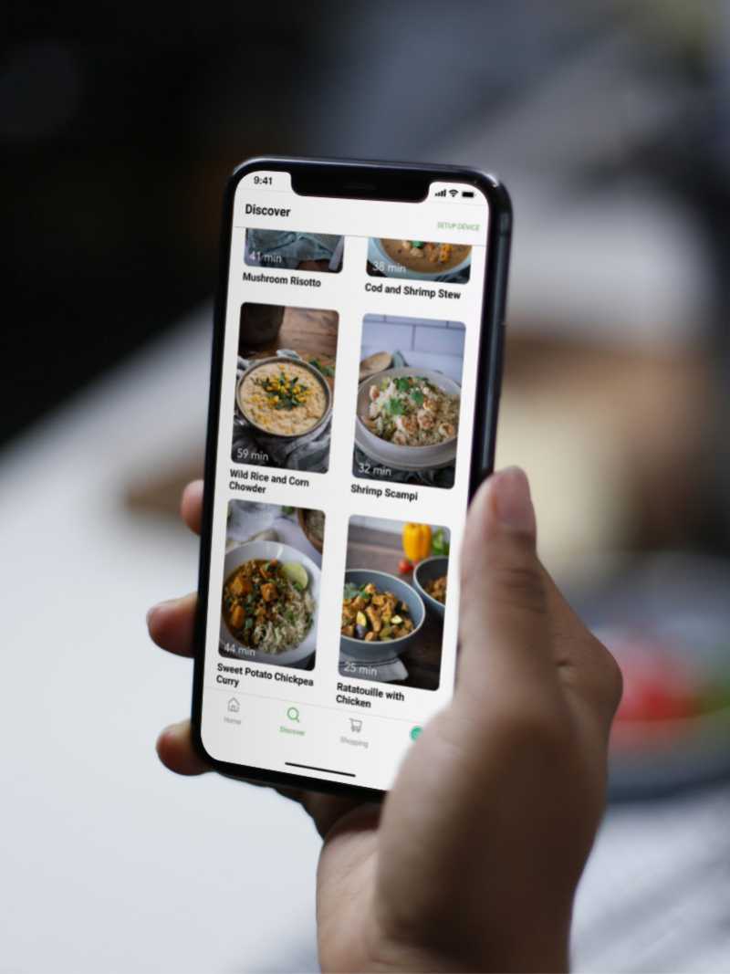 A right hand holding a phone, with the Oliver app displaying the recipe library. 
