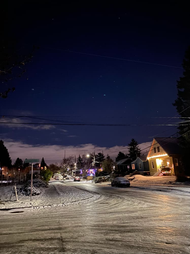 A very icy set of streets in the dark. There are a couple stars in the night sky and a couple houses brightly lit in the dark with Christmas lights.