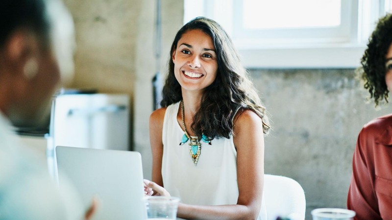 A woman smiles as she sits at a desk in front of an open laptop