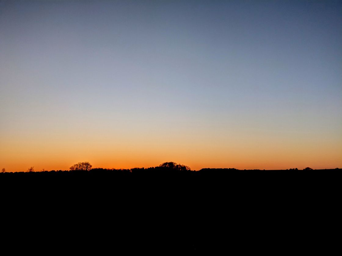 Photo of a sunset. A pitch black landscape. A few trees peak above the horizon in the far distance. The sky is a smooth cloud-less gradient from grayish blue to shiny orange at the horizon.