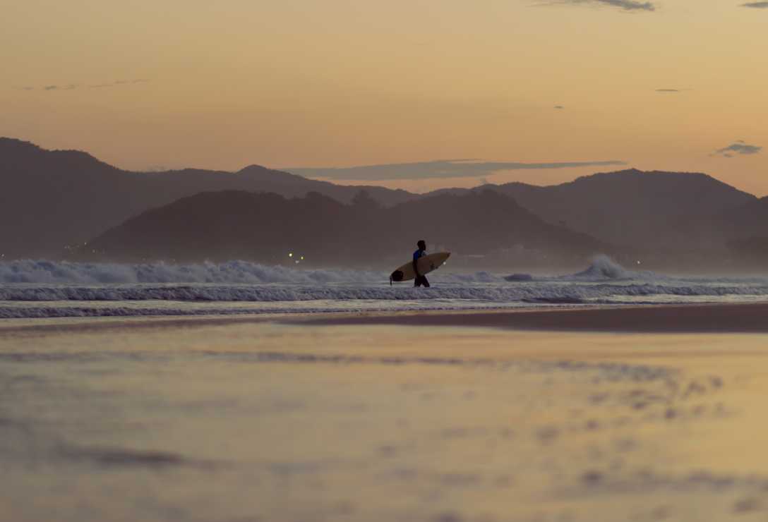 Fim de tarde com mar ao fundo e surfista saindo do mar com a prancha na mão