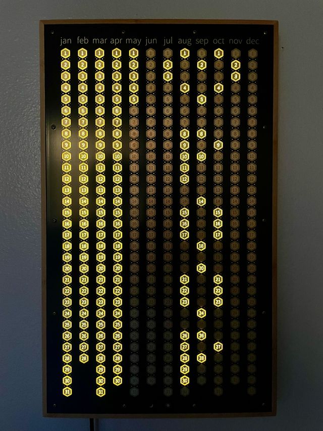 A wall-mounted wooden frame
holding a calendar with abbreviated months
along the top,
and numbered hexagon days
that light up when touched.
The first four months are fully lit,
and the rest of the days
are used to create a pixelated heart,
and spell out the word poop.
