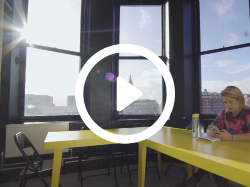 Personal meeting room containing yellow corner desk with black folding chairs in front of large windows with urban city view Skype background