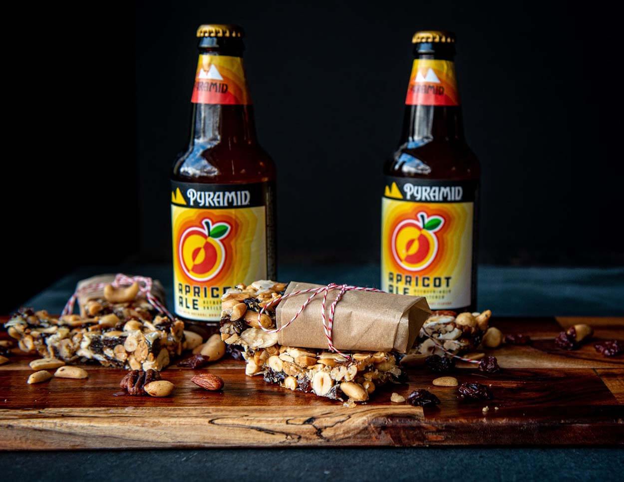 A wooden charcuterie board showing off an assortment of freshly made Apricot Ale Trail Bars along with 2 bottles of Pyramid's Apricot Ale