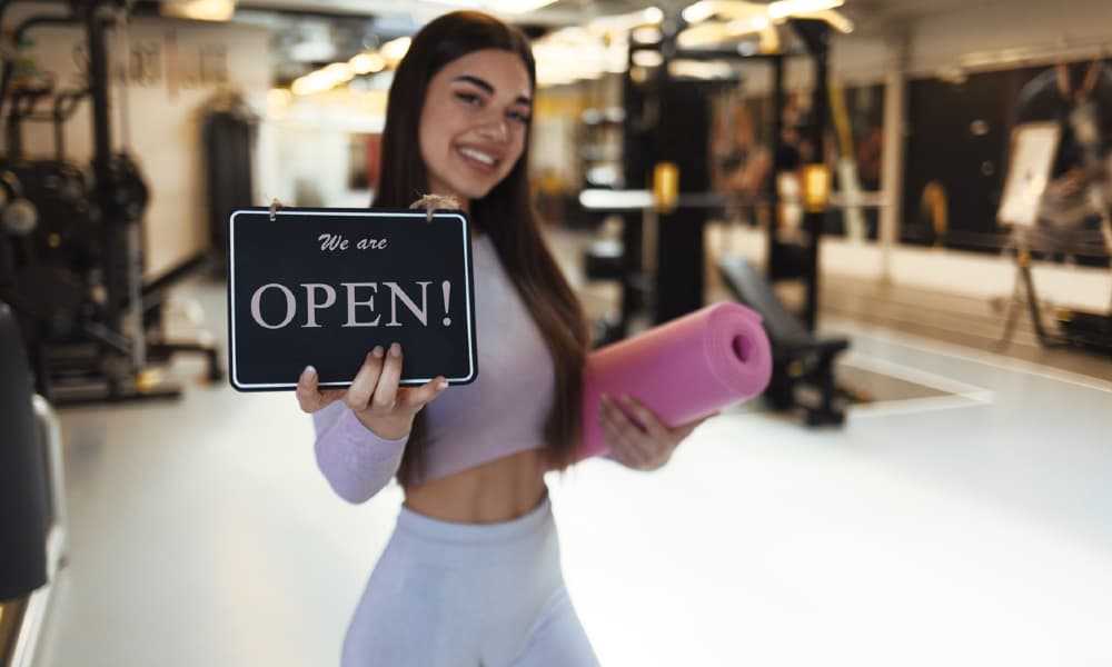 A picture of a sporty young woman holding an exercise mat and an open sign in a gym.
