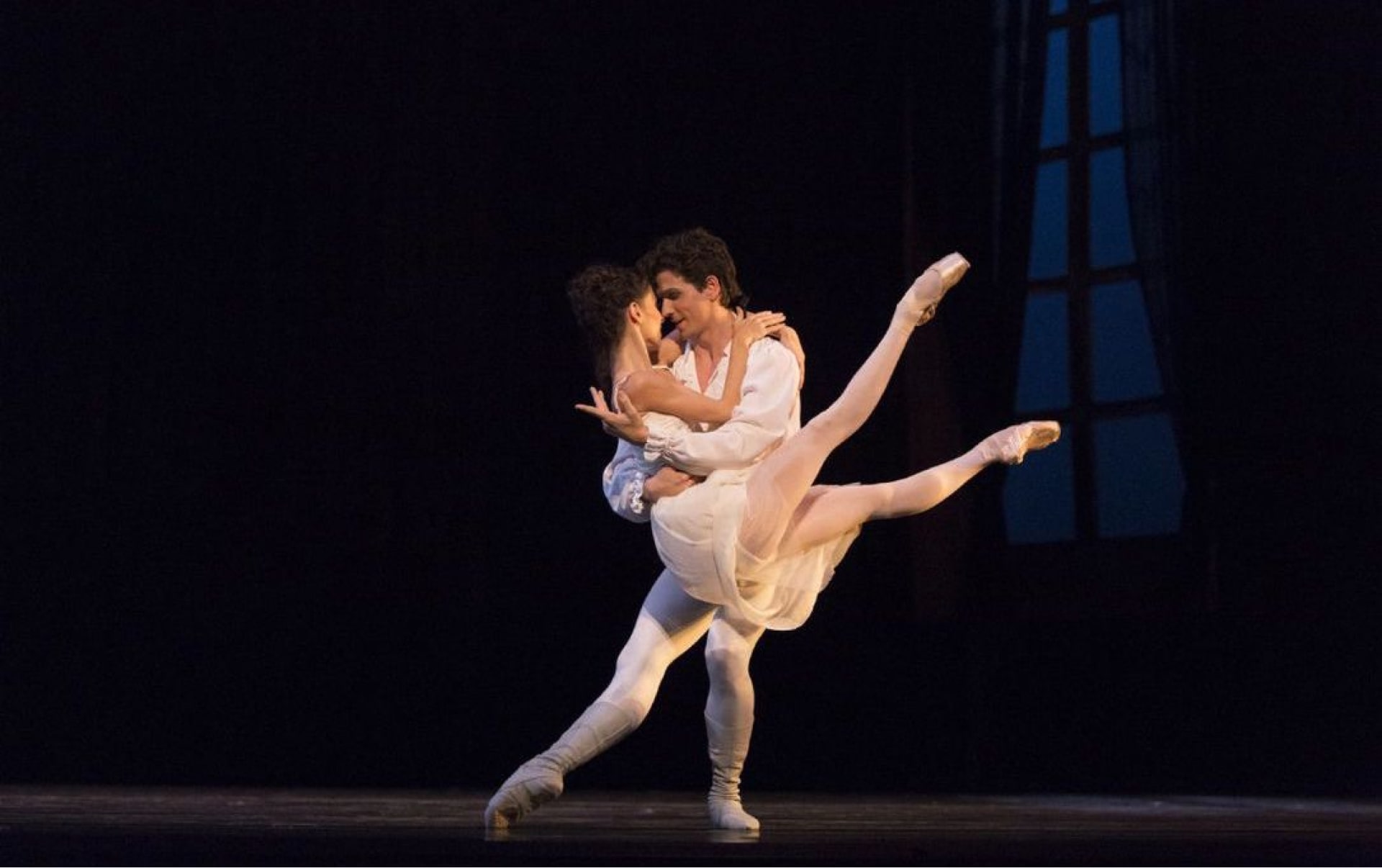 Dancer in white holds ballerina in raised embrace in front of moonlit paned window.