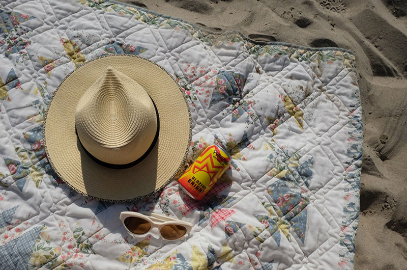 A straw hat, sunglasses, and a can of Blazing Bright on a quilted blanket on the sand