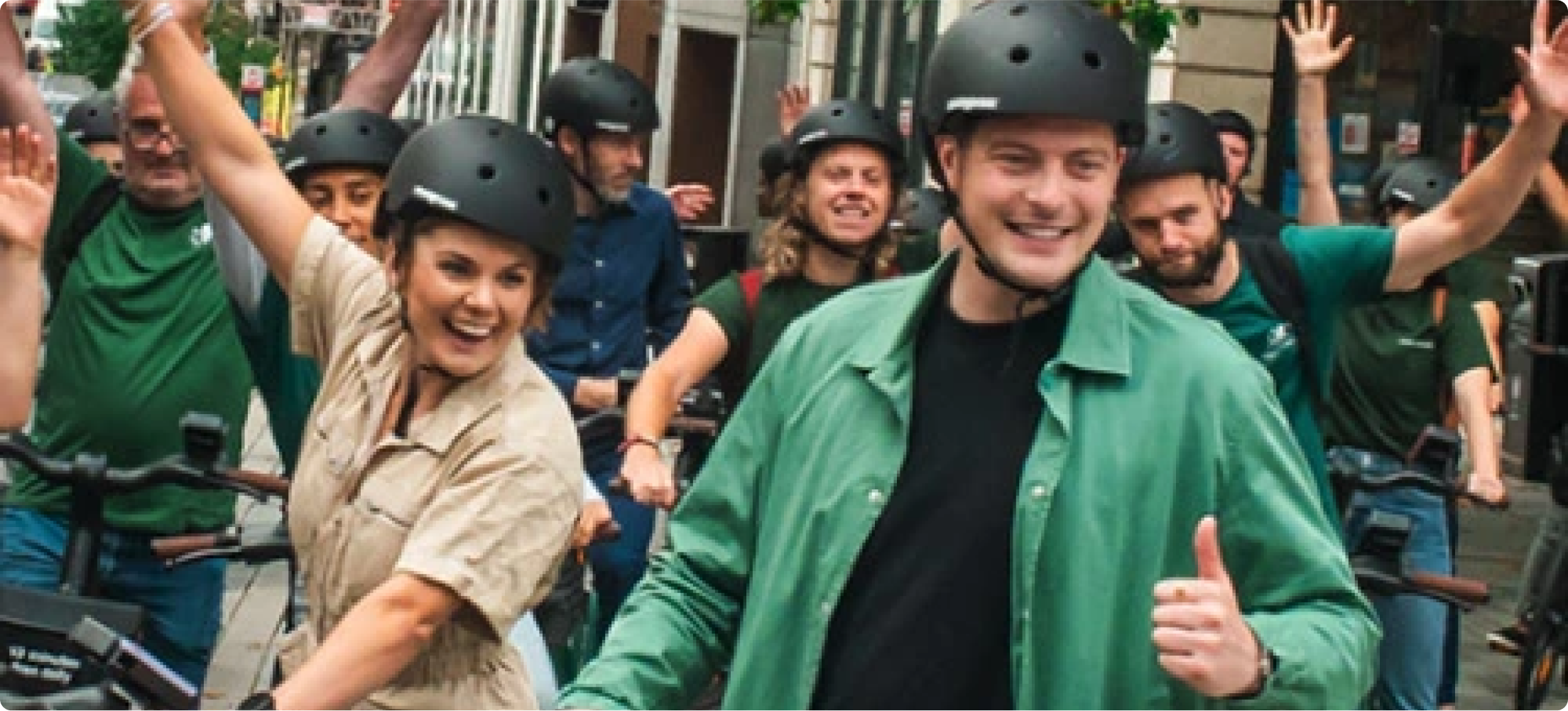 Human Forest card image with causcasian people smiling together and raising their arms while riding ebikes in an urban area.