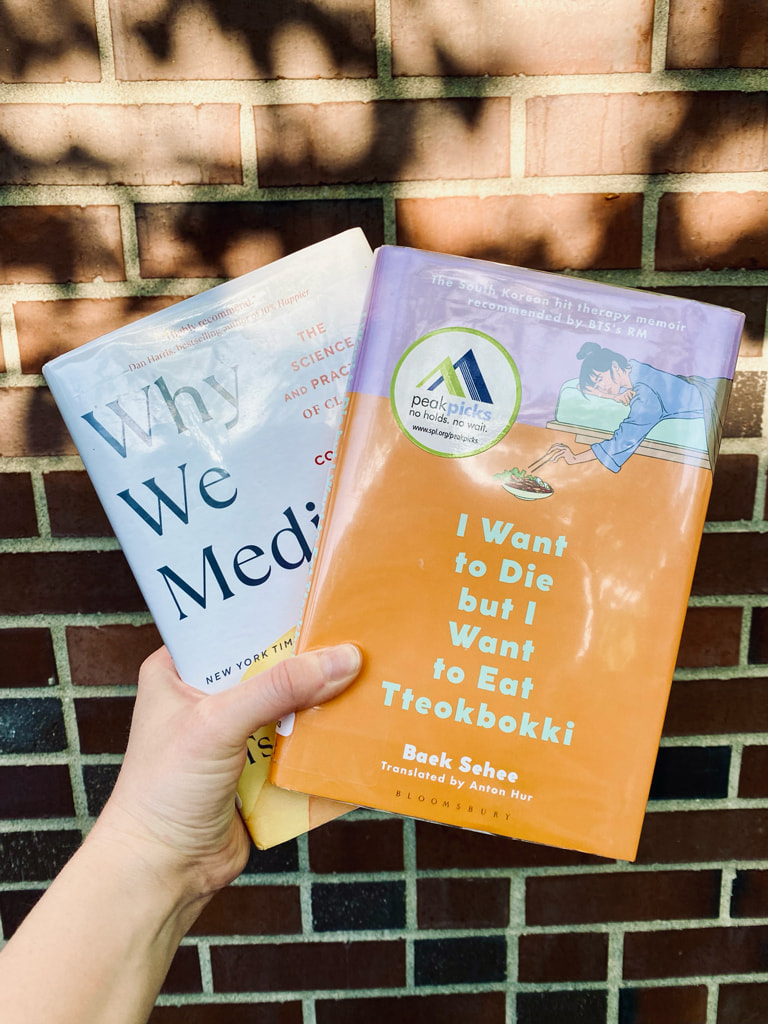 Two non-fiction books, 'Why We Medidate' and 'I Want to Die But I Want to Eat Tteokbokki, held in front of a brick wall dappled with sunlight through the trees