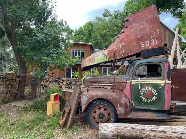 A rusting vintage pickup
with Colorado Shoe School painted on the door
and a massive metal boot mounted on the cab.
Immediately behind that,
a fence of stacked wood with ends showing,
and Erin standing behind a small metal gate under a tree.
A house and barn in the background.
