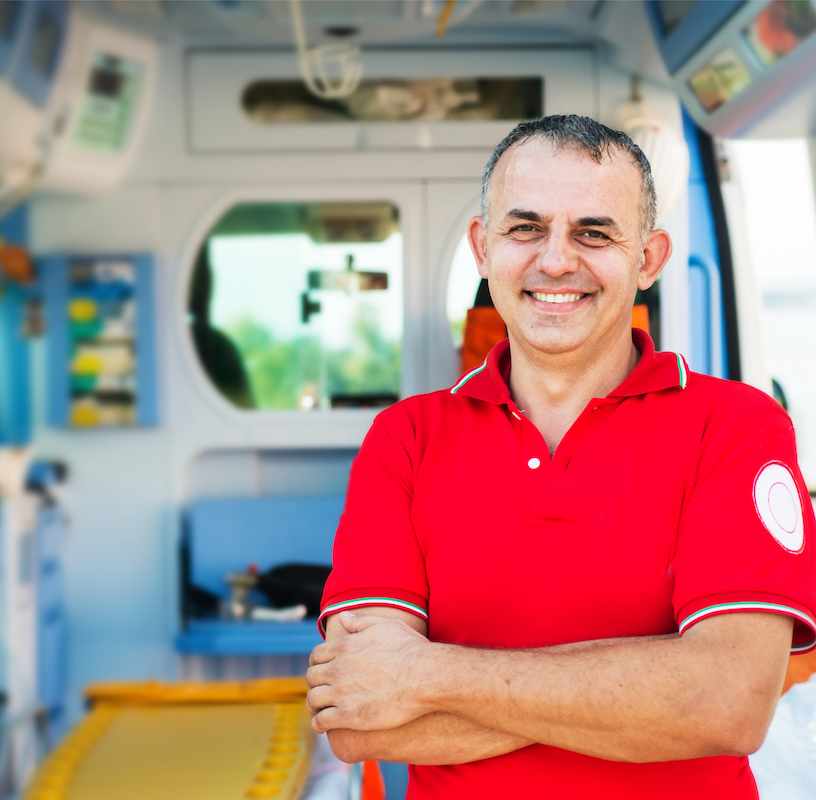 Uśmiechnięty ratownik medyczny, ubrany w czerwony uniform, stojący na tle karetki pogotowia i patrzący w obiektyw.