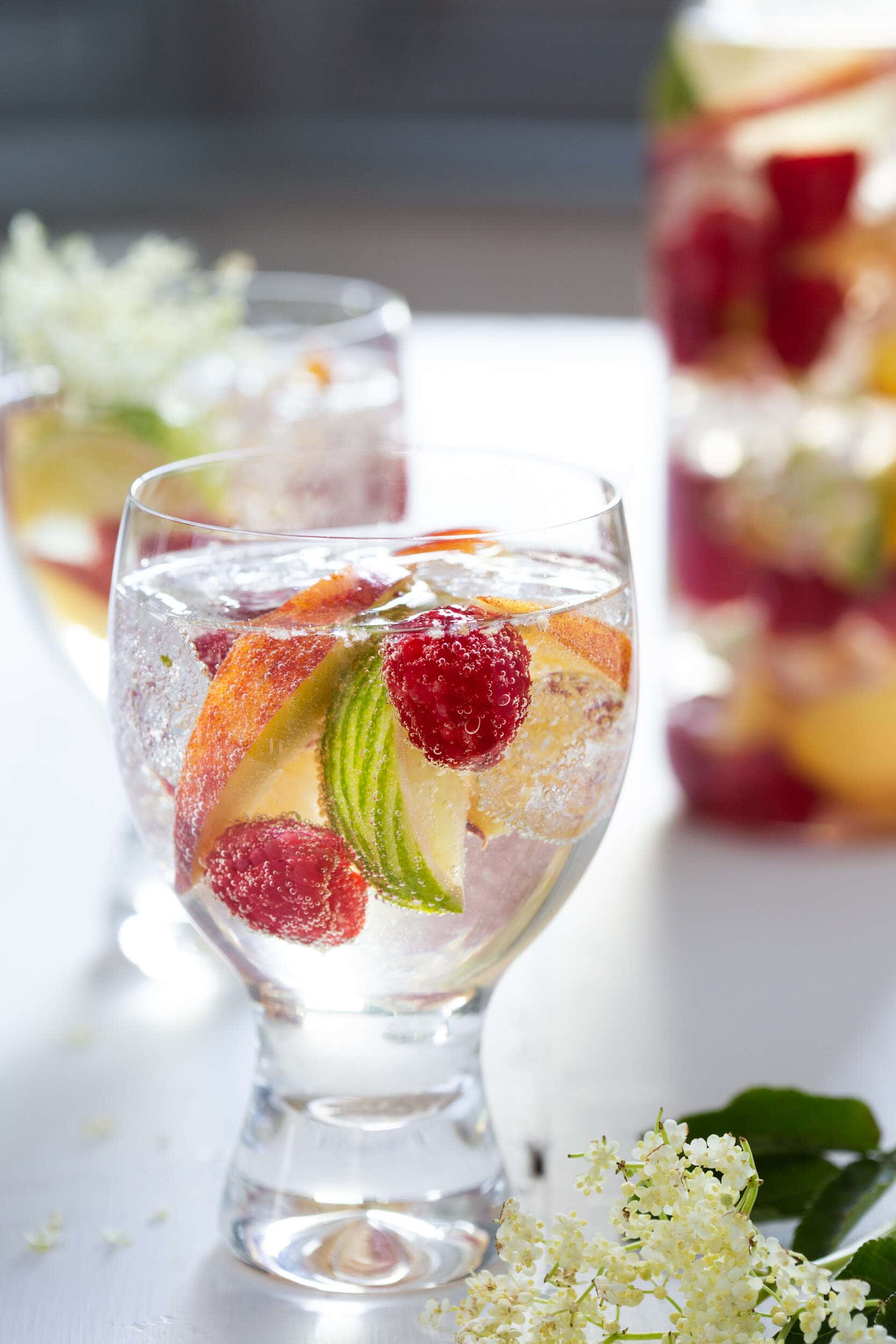White wine sangria with elderflower, raspberries, peaches and lime in a glass with a jug of the same in the background.