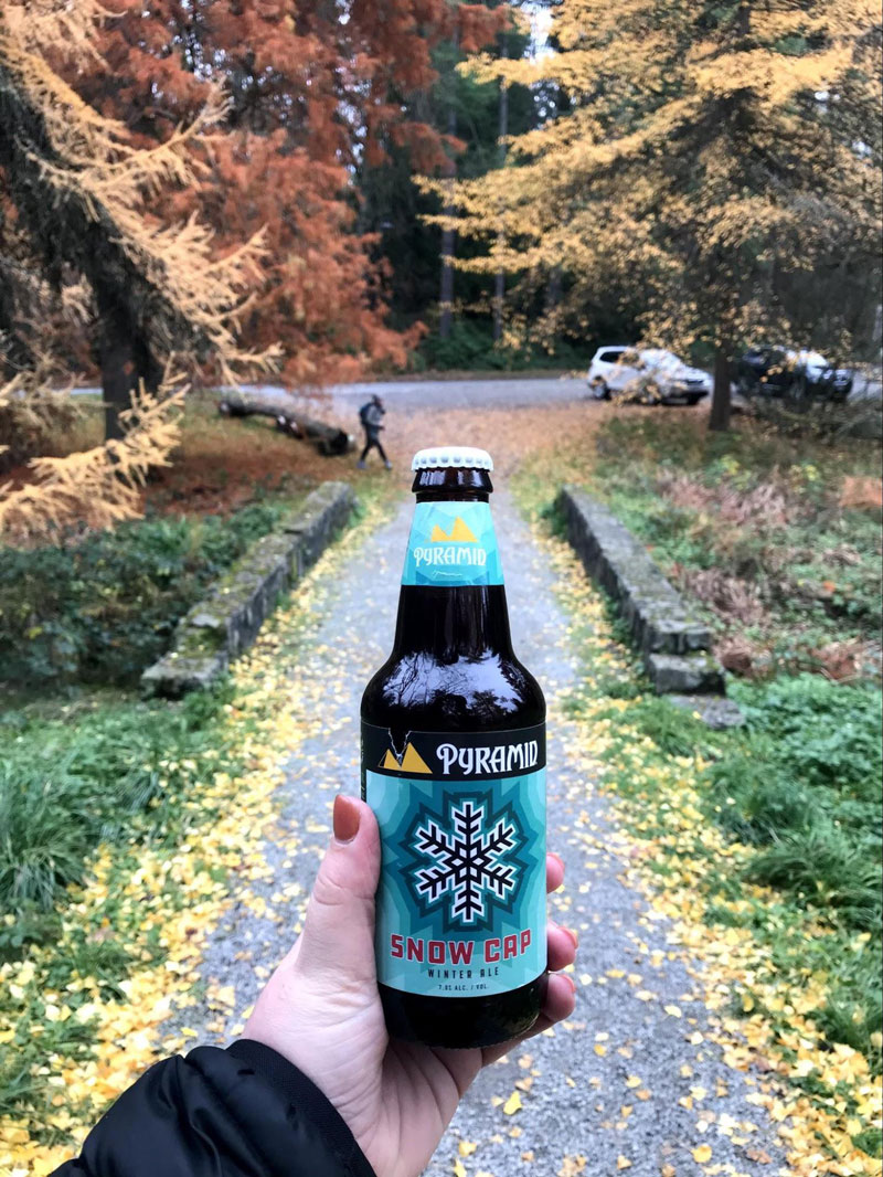 A bottle of Pyramid Snow Cap Ale in fround of a garden path covered in yellow leaves. Orange and red trees are in the background.