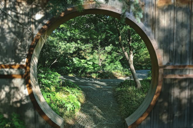 The view through a round, Chinese-style garden entrance. On the other side of the entrance, a stony path through the garden splits into two, heading off either side of the frame.