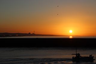 Sun rising above a harbour wall reflecting on the water. A fishing boat is in front of the harbour wall. East Sussex coastline in the distance.
