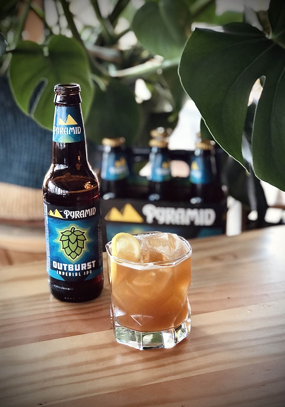 Easy recipe for summer beer cocktail with IPA and peach brandy and tea A tumbler full to the brim of the Outburst IPA beer cocktail called the Pacific Summer. The beer, peach brandy, and tea cocktail is positioned next to a bottle of Outburst IPA, a 6 pack, and some greenery.