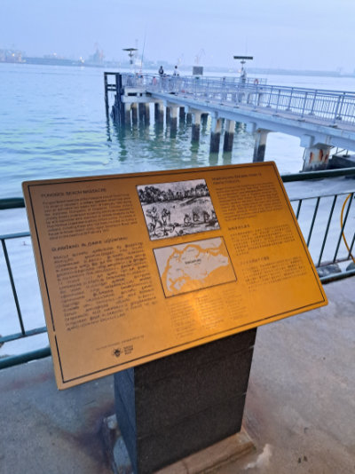A World War Two memorial plaque in the foreground, with Punggol Point Jetty and the sea in the background. The plaque details the Punggol Beach massacre.