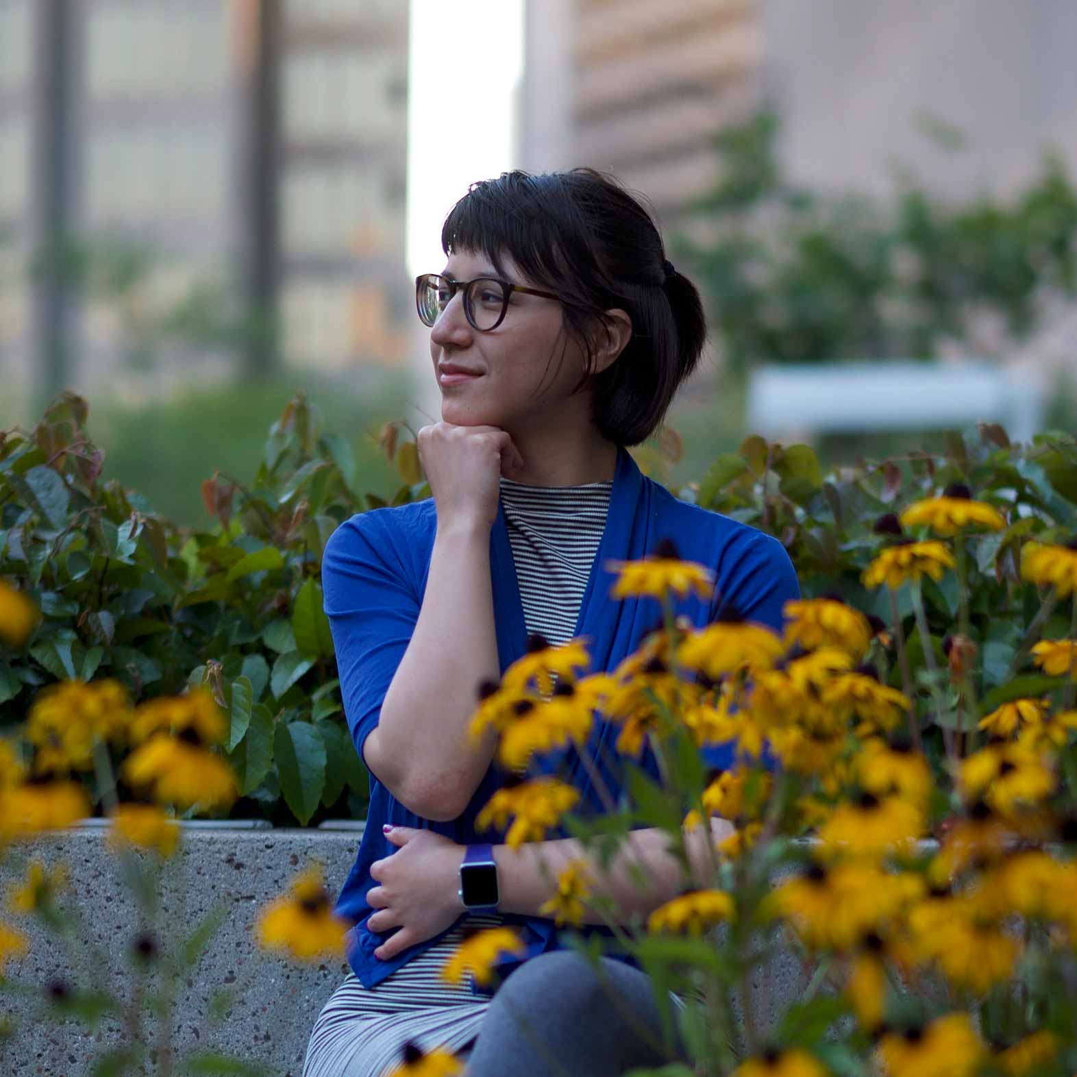 Gwen is sitting amongst flowers, her hand to her chin. She has dark hair that lays gently over her dark framed glasses. Gwen is wearing a blue cardigan over a striped shirt.
