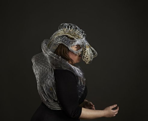 Side view of an opera singer, mid-song, while wearing the suit. This medium shot shows the tangle of tubing that wraps to the back of the head and loops around the shoulders and back—like a massive oxygen tank or backpack