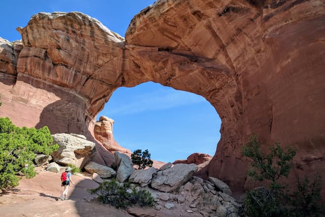 Len approaches a large bow-shaped arch made out of red sandstone. White sandstone boulders, many larger than her, litter its base.