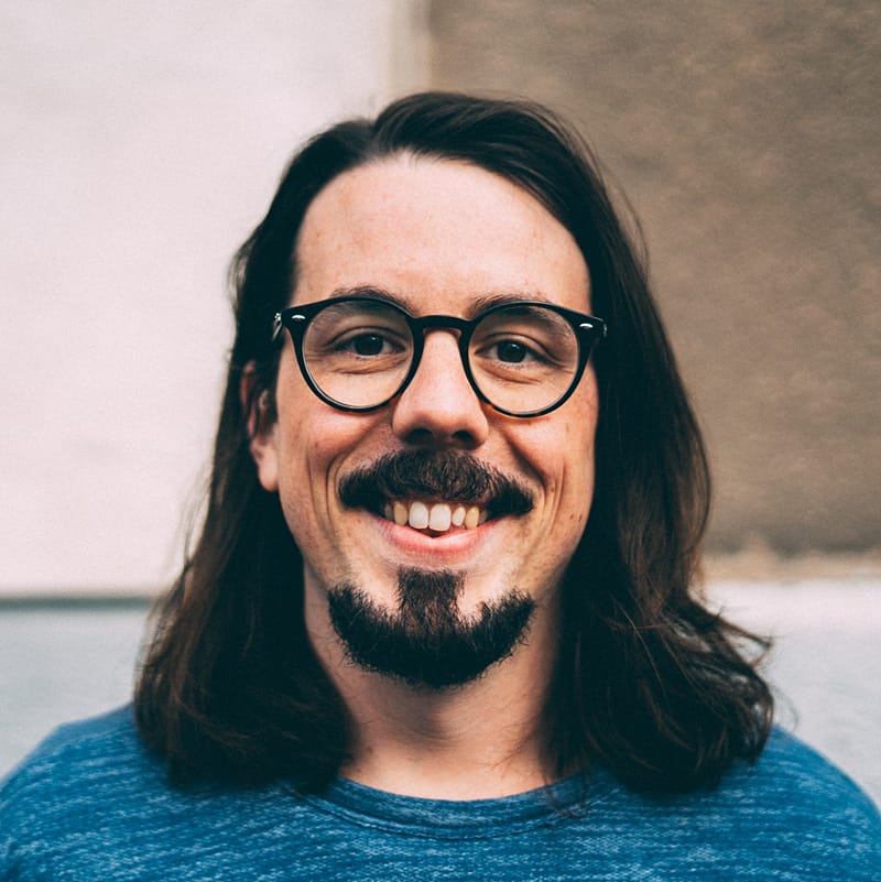 Portrait of a smiling, bearded, and long-haired man wearing glasses.