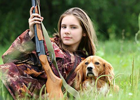 A young girl lays on the grass with a gun in her hand and a dog next to her. Taking kids hunting is great for them.