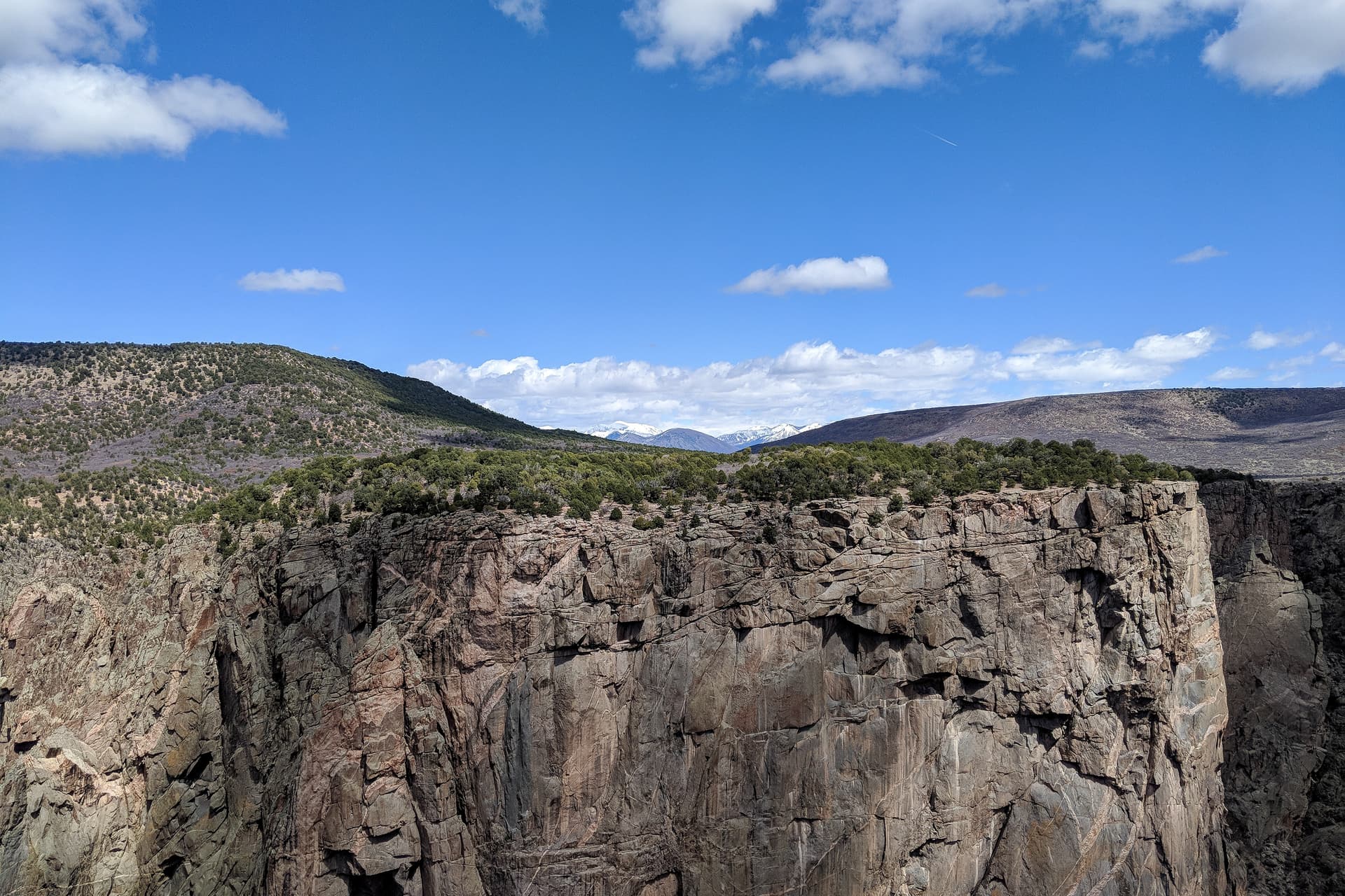 Looking straight across the Black Canyon of the Gunnison to the north rim's high plateau. In the distance, ramed by two low, wide hills, can be seen a range of domed, snow-capped mountains.