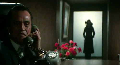 A screenshot from the film 'Female Prisoner 701: Scorpion' of a older man on the telephone oblivious to the silhouette of a woman with a large hat behind him in the distance.