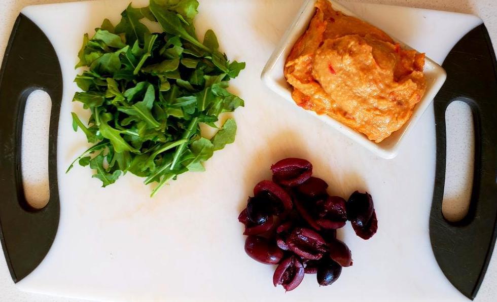 Cutting board with olives, arugula, red pepper hummus