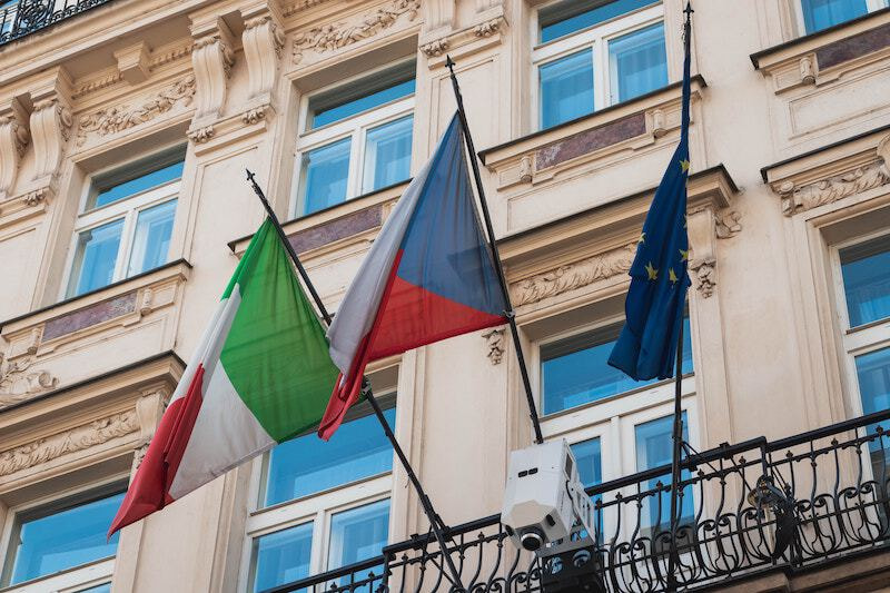 Different EU flags hanging on a balcony of a renaissance inspired building.