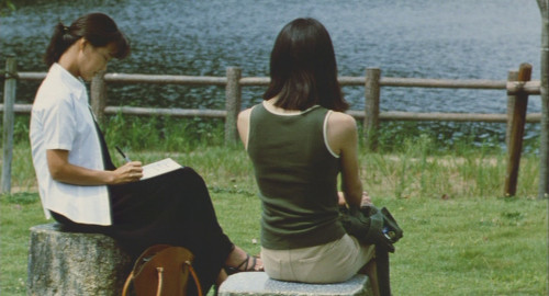 A screenshot from the movie 'Tomie' of a young woman, Tsukiko, sitting outside with her back to the camera talking to her therapist.