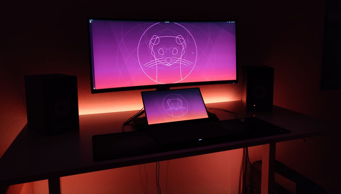 Orange illuminated desk with ultra-wide monitor and notebook showing the Ubuntu 19.10 “Eoan Ermine” wallpaper.
