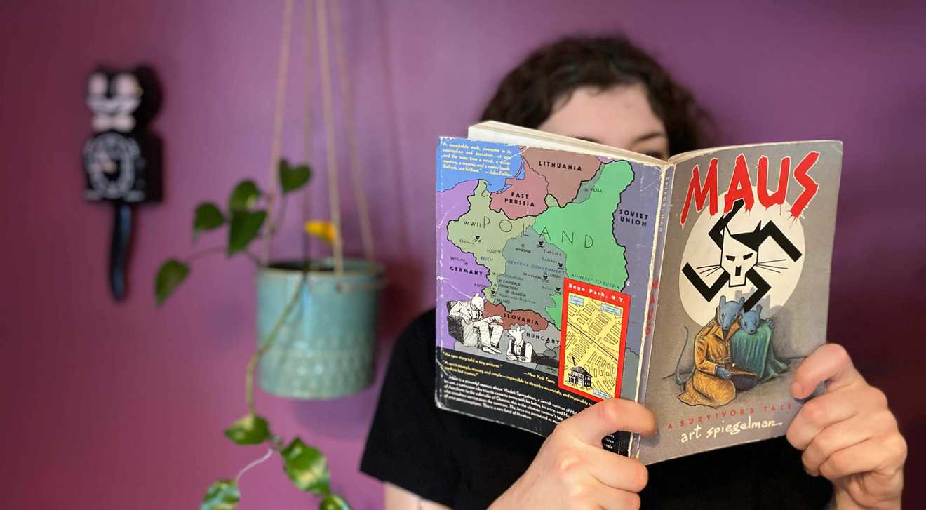 Photo of a young woman reading the book Maus with a colourful background with a plant and a cat clock over her shoulder