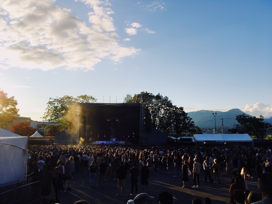 An outdoor concert with a crowd standing in front of the stage. In the background are mountains and clouds, and the sun is getting low.