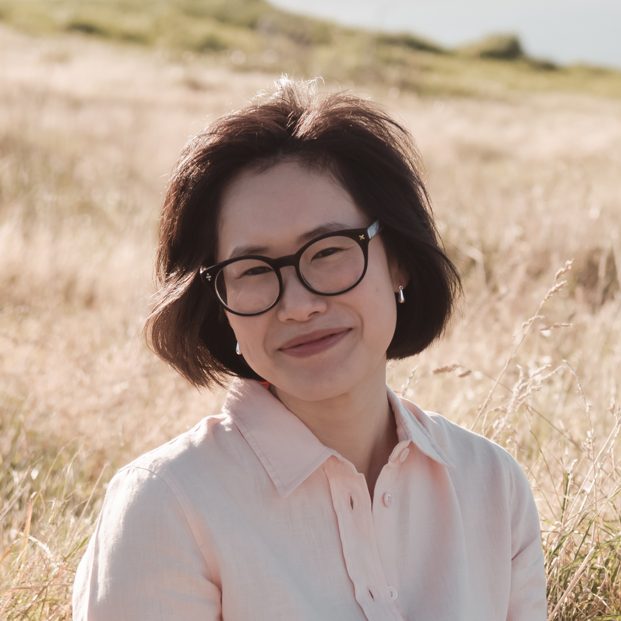 Headshot of Prae Songprasit with semi-long black hair, black glasses, smiling kindly at the camera. She's wearing a light peach shirt, with golden grassy background