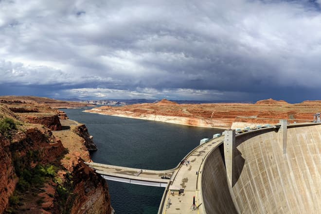 The Glen Canyon Dam, with Lake Powell extending behind it to the horizon. The rocks on either side of the lake are bright red-orange, but turn white just above the water. A thunder storm can be seen in the distance.