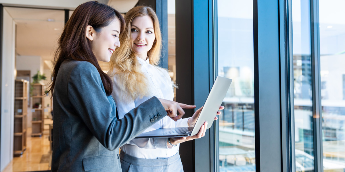 Data analytics job descriptions and what they really mean Two women smiling at a laptop screen, standing by a glass window