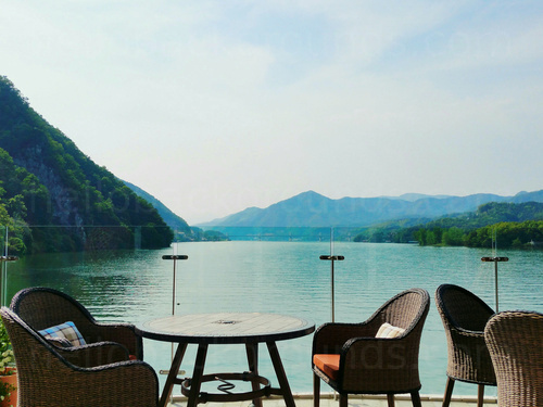 Scenic outdoor cafe deck with round wooden table and wicker chairs in front of mountain lake view Microsoft Teams background