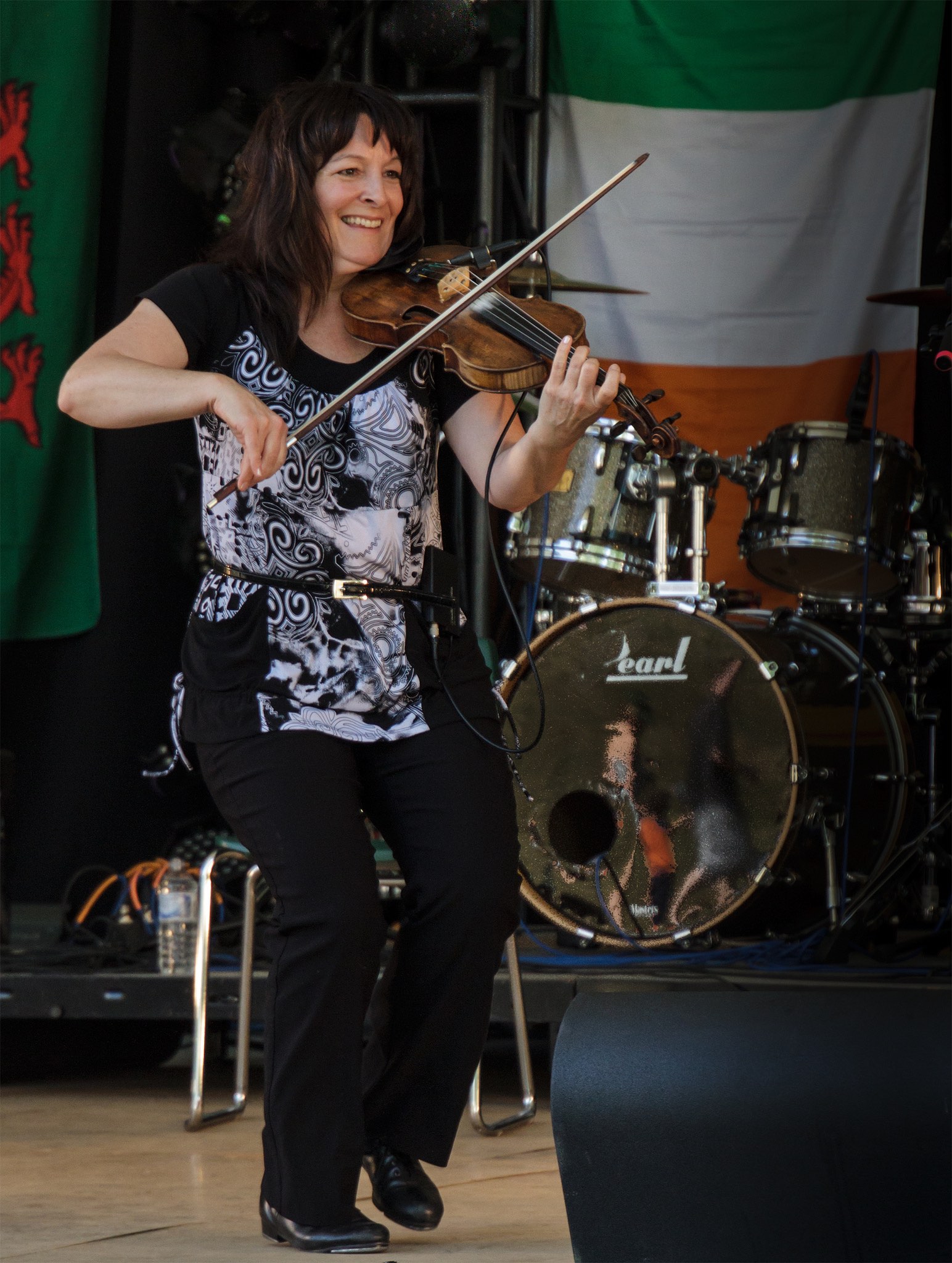 Cindy Thompson similing, step dancing and playing the fiddle on stage in front of a set of drums.