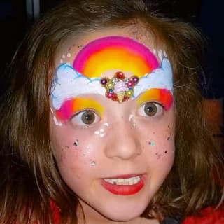A rainbow and fluffy clouds are painted on this girl's forehead. Some colourful ornaments are attached above her nose with adhesive, and some glitter adds a sparkle to her cheeks. Click to view at full size.