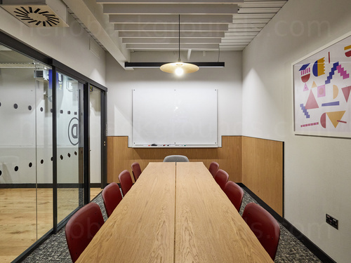 Simple board room with large interior windows containing basic wooden table and red fabric chairs beside whiteboard  Microsoft Teams background