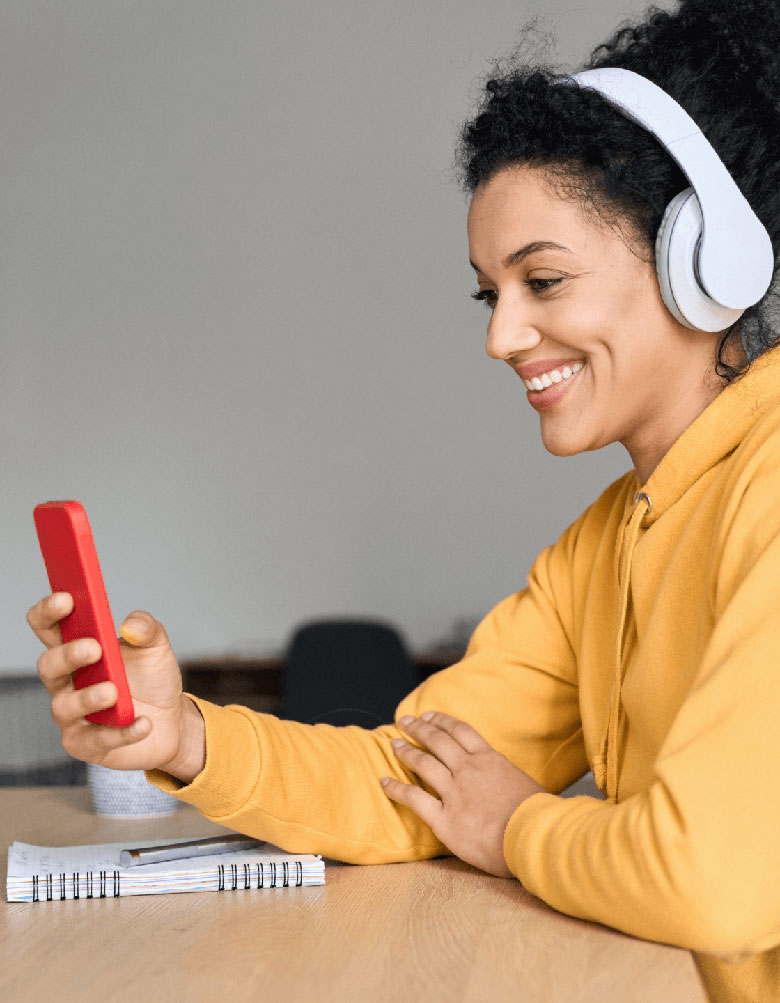 Image of a woman at home wearing headphones and smiling at her phone screen.