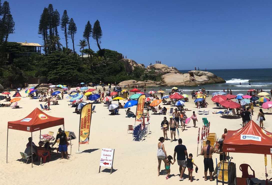 Vista da area com banhistas e guarda-sóis. Mar ao fundo com um pouco de vegetação e pedras.