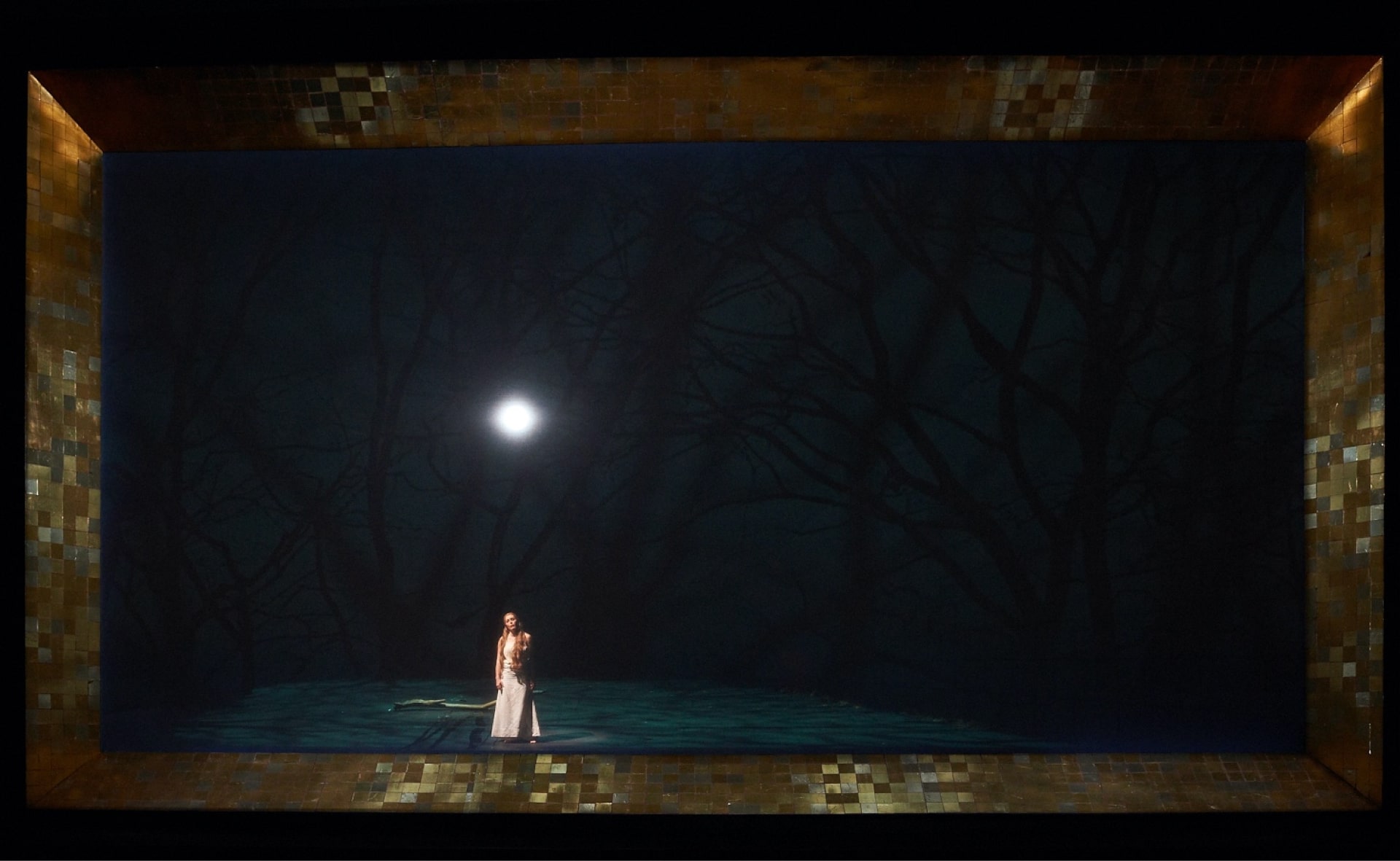 Woman in white stands in projected green forest under gleaming moon, behind gold-tiled frame.