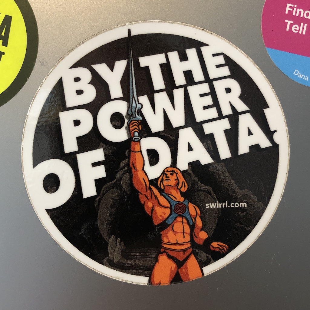 A round sticker that says 'by the power of data'. He-Man stands in front, raising his sword.