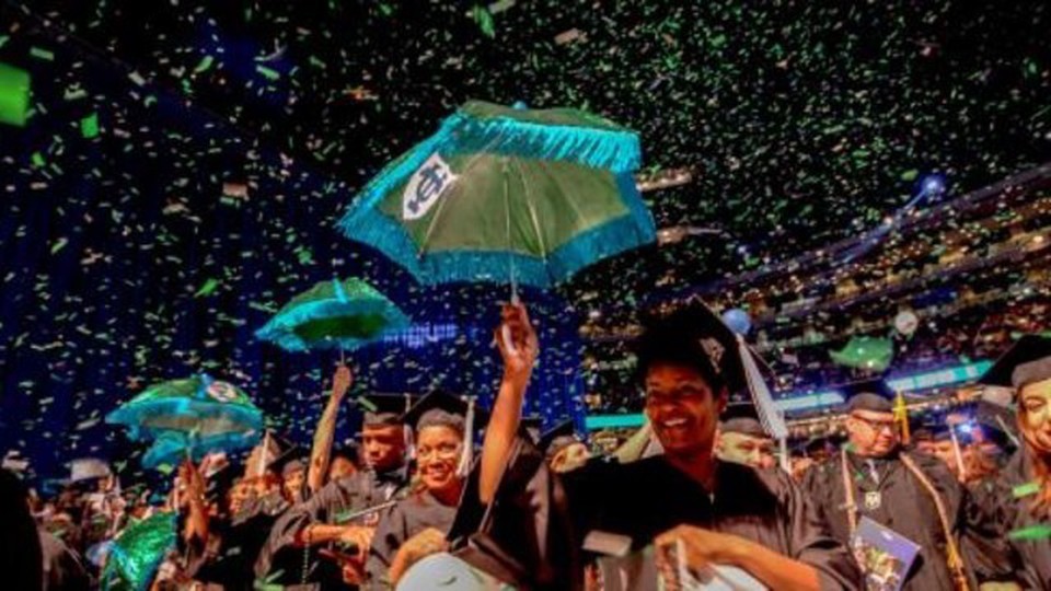 A line of people holding a umbrella with Tulane University's logo dressed with a graduation cap. There's paper falling all over them.
